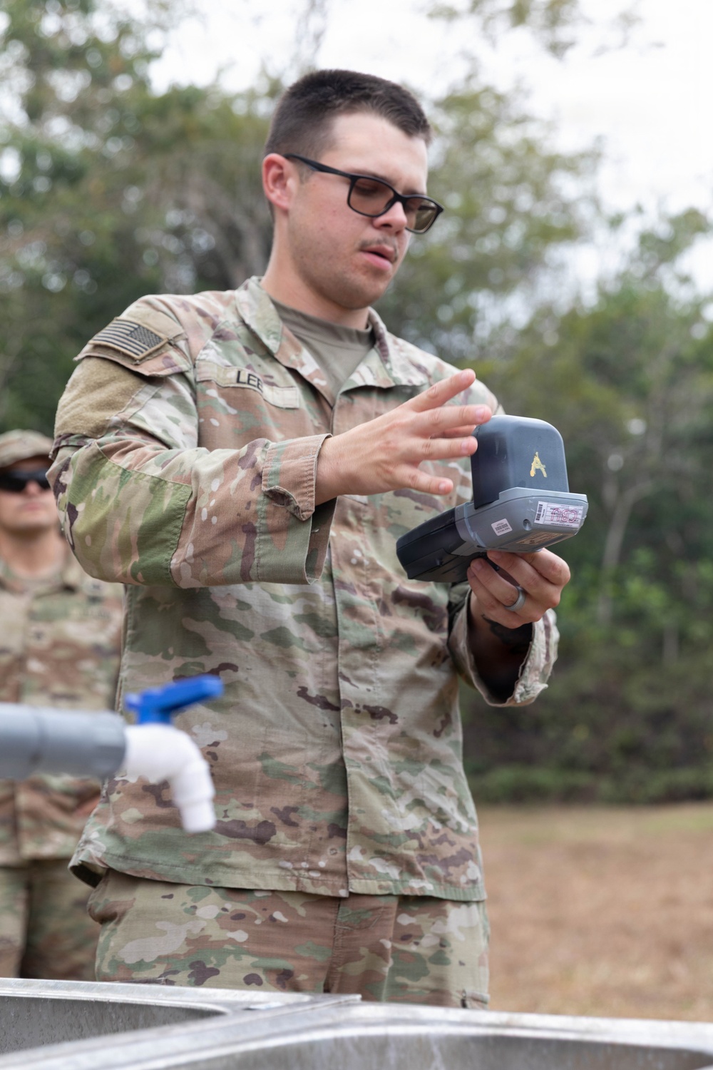 A U.S. Army soldier wearing OCP (Operational Camouflage Pattern) uniform is holding and examining a handheld electronic device, likely a water testing or detection instrument. The soldier, whose name tape reads 'LEE', appears to be conducting a field demonstration or training exercise near what appears to be a water purification or field sanitation station. A second soldier is partially visible in the background.