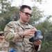A U.S. Army soldier wearing OCP (Operational Camouflage Pattern) uniform is holding and examining a handheld electronic device, likely a water testing or detection instrument. The soldier, whose name tape reads 'LEE', appears to be conducting a field demonstration or training exercise near what appears to be a water purification or field sanitation station. A second soldier is partially visible in the background.