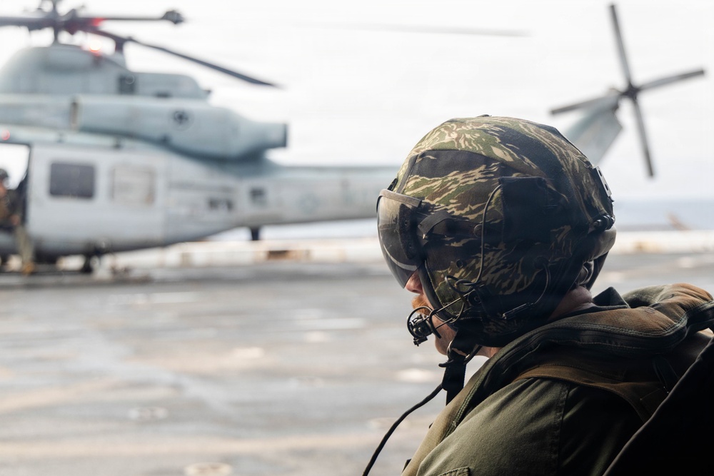 A military personnel wearing a camouflage flight helmet with visor and communications equipment observes a large military helicopter on what appears to be a ship's flight deck. The helicopter in the background is a large tandem-rotor or heavy-lift type aircraft, consistent with a CH-46 or CH-53 series. The scene suggests a naval aviation or amphibious operation environment.