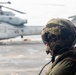 A military personnel wearing a camouflage flight helmet with visor and communications equipment observes a large military helicopter on what appears to be a ship's flight deck. The helicopter in the background is a large tandem-rotor or heavy-lift type aircraft, consistent with a CH-46 or CH-53 series. The scene suggests a naval aviation or amphibious operation environment.