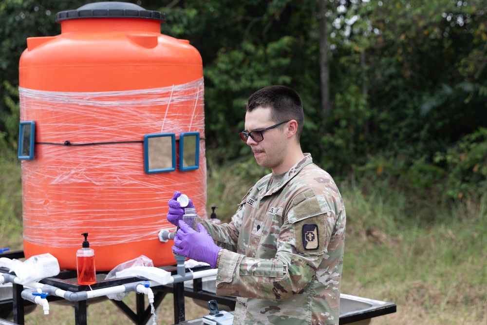 A U.S. Army soldier in OCP camouflage uniform is conducting water testing or sampling operations outdoors. He is wearing purple nitrile gloves and handling a sample container near a large orange water storage tank wrapped in plastic sheeting. The setting appears to be a field environment with trees in the background, suggesting a field training exercise or disaster relief water purification operation.
