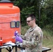 A U.S. Army soldier in OCP camouflage uniform is conducting water testing or sampling operations outdoors. He is wearing purple nitrile gloves and handling a sample container near a large orange water storage tank wrapped in plastic sheeting. The setting appears to be a field environment with trees in the background, suggesting a field training exercise or disaster relief water purification operation.