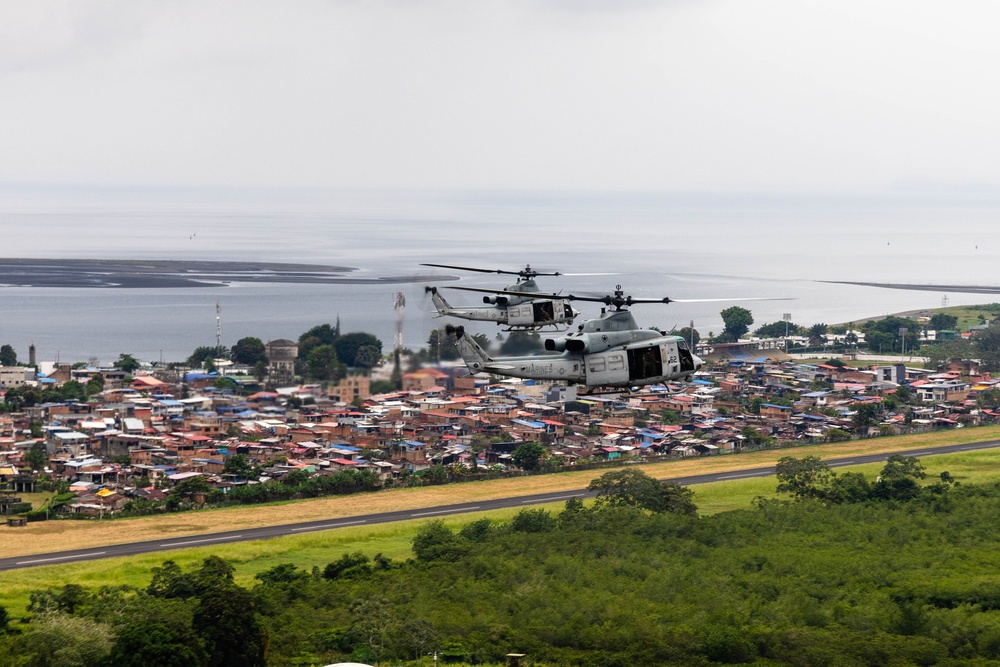 Two military helicopters, identifiable as U.S. Marines aircraft, are flying in formation low over a runway adjacent to a densely populated coastal town. The scene shows a tropical urban area with colorful rooftops, lush vegetation, and a body of water visible in the background. The helicopters appear to be on an approach or departure path near what looks like a small regional airport.