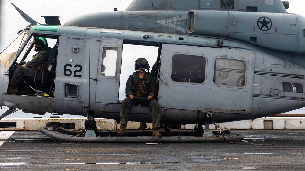 A U.S. military helicopter, numbered '62', is parked on what appears to be a naval vessel's flight deck. Two military personnel in flight gear and helmets are visible — one seated in the cockpit and one sitting in the open side door. The aircraft bears U.S. military markings including a star insignia, and the scene appears to be taken in overcast, wet conditions at sea.