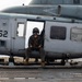 A U.S. military helicopter, numbered '62', is parked on what appears to be a naval vessel's flight deck. Two military personnel in flight gear and helmets are visible — one seated in the cockpit and one sitting in the open side door. The aircraft bears U.S. military markings including a star insignia, and the scene appears to be taken in overcast, wet conditions at sea.