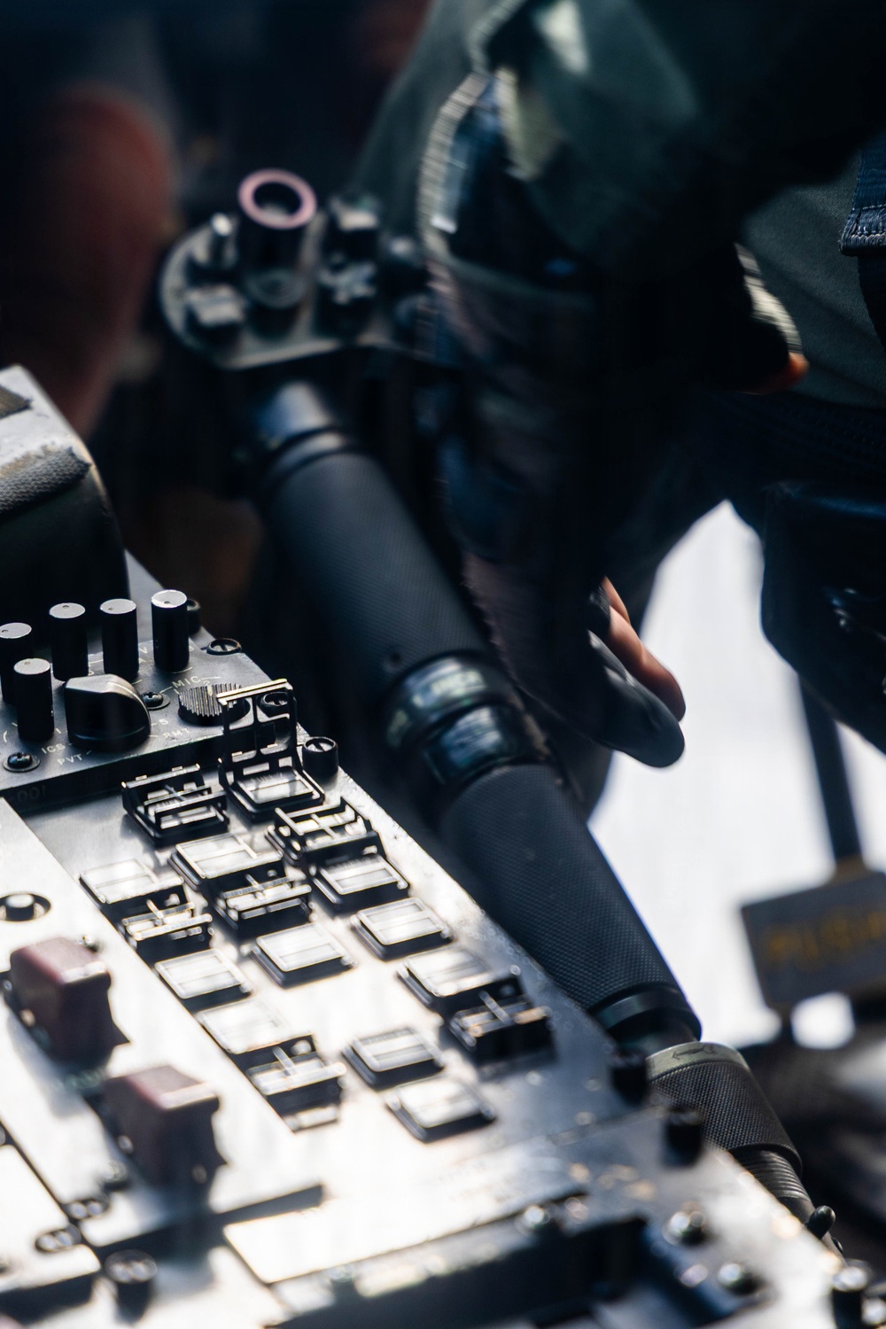 A close-up photograph of what appears to be an aircraft cockpit control panel, featuring a throttle or control stick being gripped by a gloved hand. The panel contains numerous toggle switches, knobs, and buttons typical of military or commercial aviation instrumentation. The image is shot with shallow depth of field, keeping the foreground controls in sharp focus while the background is blurred.