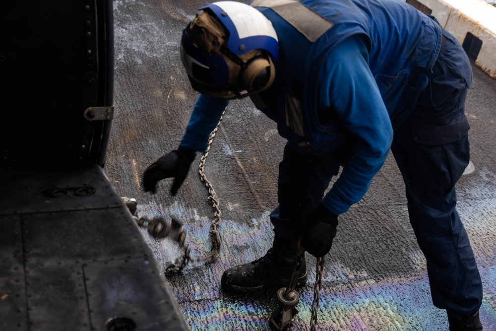 A naval deck crew member dressed in blue coveralls and a blue helmet with hearing protection is handling tie-down chains on what appears to be an aircraft carrier flight deck. The deck surface shows iridescent oil sheen patterns, indicating a wet or fuel-contaminated surface. The worker is bent over, managing metal chains used to secure aircraft to the deck.