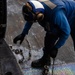 A naval deck crew member dressed in blue coveralls and a blue helmet with hearing protection is handling tie-down chains on what appears to be an aircraft carrier flight deck. The deck surface shows iridescent oil sheen patterns, indicating a wet or fuel-contaminated surface. The worker is bent over, managing metal chains used to secure aircraft to the deck.