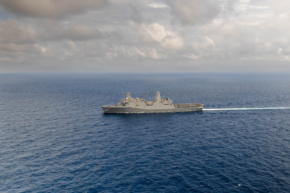 An aerial photograph of a large gray naval warship underway in open blue ocean waters. The vessel appears to be a U.S. Navy amphibious transport dock (LPD-class) ship, identifiable by its flat flight deck, superstructure configuration, and gray hull. The ship is leaving a visible white wake as it travels, photographed under a partly cloudy sky.