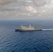 An aerial photograph of a large gray naval warship underway in open blue ocean waters. The vessel appears to be a U.S. Navy amphibious transport dock (LPD-class) ship, identifiable by its flat flight deck, superstructure configuration, and gray hull. The ship is leaving a visible white wake as it travels, photographed under a partly cloudy sky.