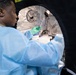 A laboratory technician wearing blue PPE gown and green nitrile gloves is working inside what appears to be a portable biosafety containment tent or enclosure. The worker is handling a small red-capped specimen vial or tube near laboratory equipment including what appears to be a centrifuge. The setting suggests a field laboratory or mobile diagnostic unit operating under biosafety protocols.