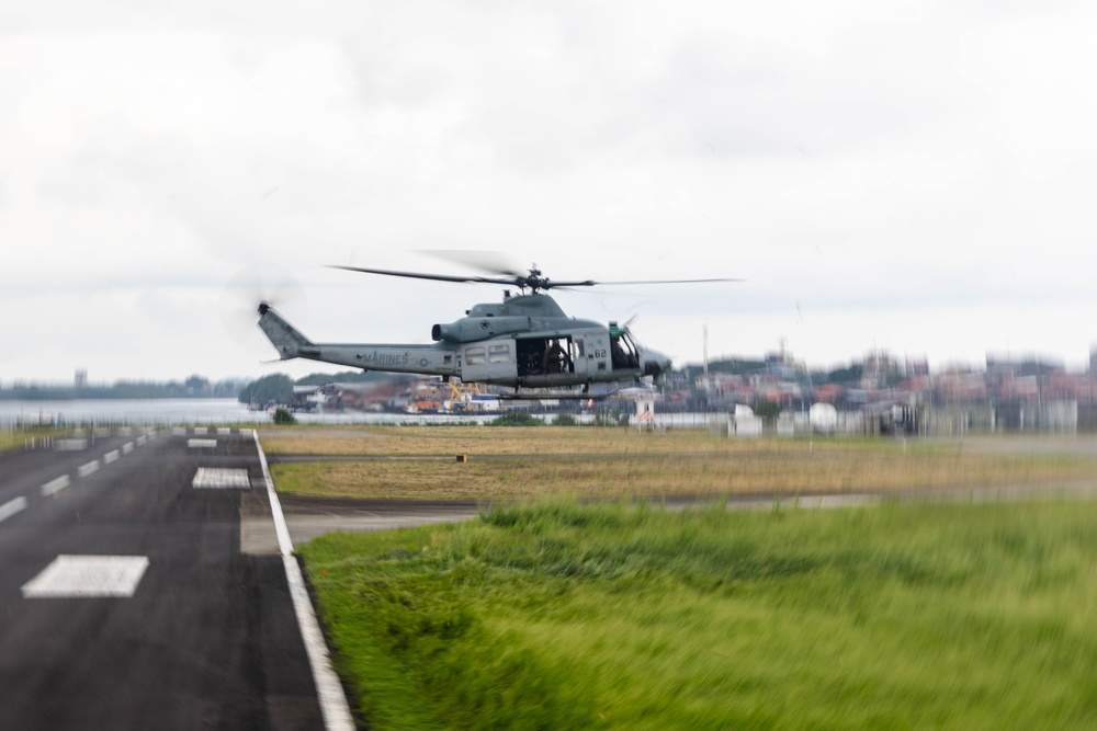 A U.S. Marines military helicopter, appearing to be a UH-1Y Venom or similar Bell helicopter variant, is captured in low-altitude flight near an airfield runway. The aircraft bears 'MARINES' markings and the tail number '02', flying just above the ground in what appears to be a takeoff or landing approach. The background shows an industrial or port area with structures visible through the overcast conditions.