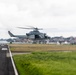 A U.S. Marines military helicopter, appearing to be a UH-1Y Venom or similar Bell helicopter variant, is captured in low-altitude flight near an airfield runway. The aircraft bears 'MARINES' markings and the tail number '02', flying just above the ground in what appears to be a takeoff or landing approach. The background shows an industrial or port area with structures visible through the overcast conditions.