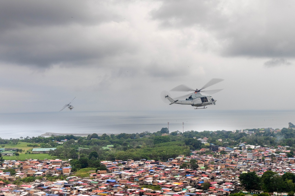 Two military-style helicopters are flying over a densely populated coastal urban area under overcast skies. The foreground helicopter is clearly visible and appears to be a light utility helicopter, while a second aircraft is visible in the background to the left. The city below features tightly packed, colorful low-rise buildings extending toward a coastline with visible water and green vegetation.
