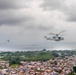 Two military-style helicopters are flying over a densely populated coastal urban area under overcast skies. The foreground helicopter is clearly visible and appears to be a light utility helicopter, while a second aircraft is visible in the background to the left. The city below features tightly packed, colorful low-rise buildings extending toward a coastline with visible water and green vegetation.