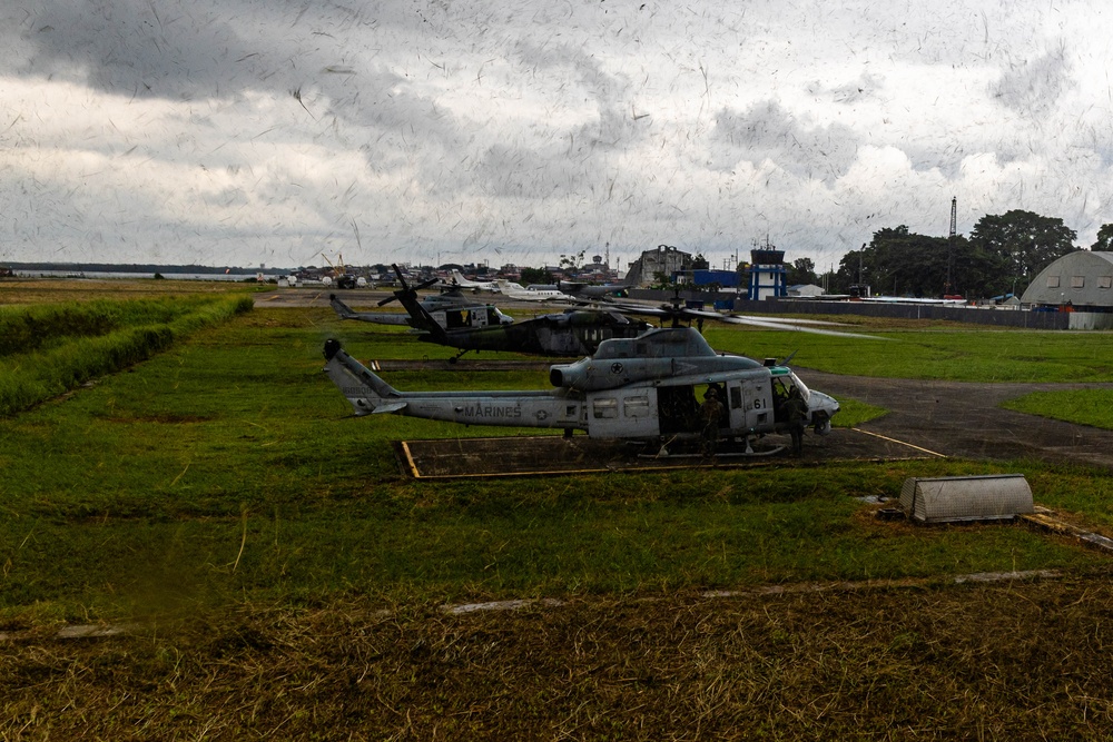 Multiple military helicopters are parked on a grass airfield, with the foreground featuring a U.S. Marines-marked helicopter (likely a UH-1Y Venom or similar Bell variant) bearing the number '61'. Behind it are additional darker helicopters, possibly UH-60 Black Hawks. The setting appears to be a small regional airfield with a control tower, hangars, and civilian aircraft visible in the background under overcast skies.