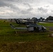 Multiple military helicopters are parked on a grass airfield, with the foreground featuring a U.S. Marines-marked helicopter (likely a UH-1Y Venom or similar Bell variant) bearing the number '61'. Behind it are additional darker helicopters, possibly UH-60 Black Hawks. The setting appears to be a small regional airfield with a control tower, hangars, and civilian aircraft visible in the background under overcast skies.