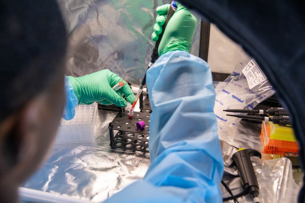Laboratory workers wearing blue protective gowns and green nitrile gloves are handling blood collection tubes and laboratory equipment inside what appears to be a biosafety containment enclosure with a clear plastic barrier. The workspace contains multiple scientific instruments and supplies including Eppendorf tubes, pipettes, and a tube rack. The setting suggests a high-containment or biosafety-level laboratory procedure.