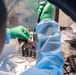 Laboratory workers wearing blue protective gowns and green nitrile gloves are handling blood collection tubes and laboratory equipment inside what appears to be a biosafety containment enclosure with a clear plastic barrier. The workspace contains multiple scientific instruments and supplies including Eppendorf tubes, pipettes, and a tube rack. The setting suggests a high-containment or biosafety-level laboratory procedure.