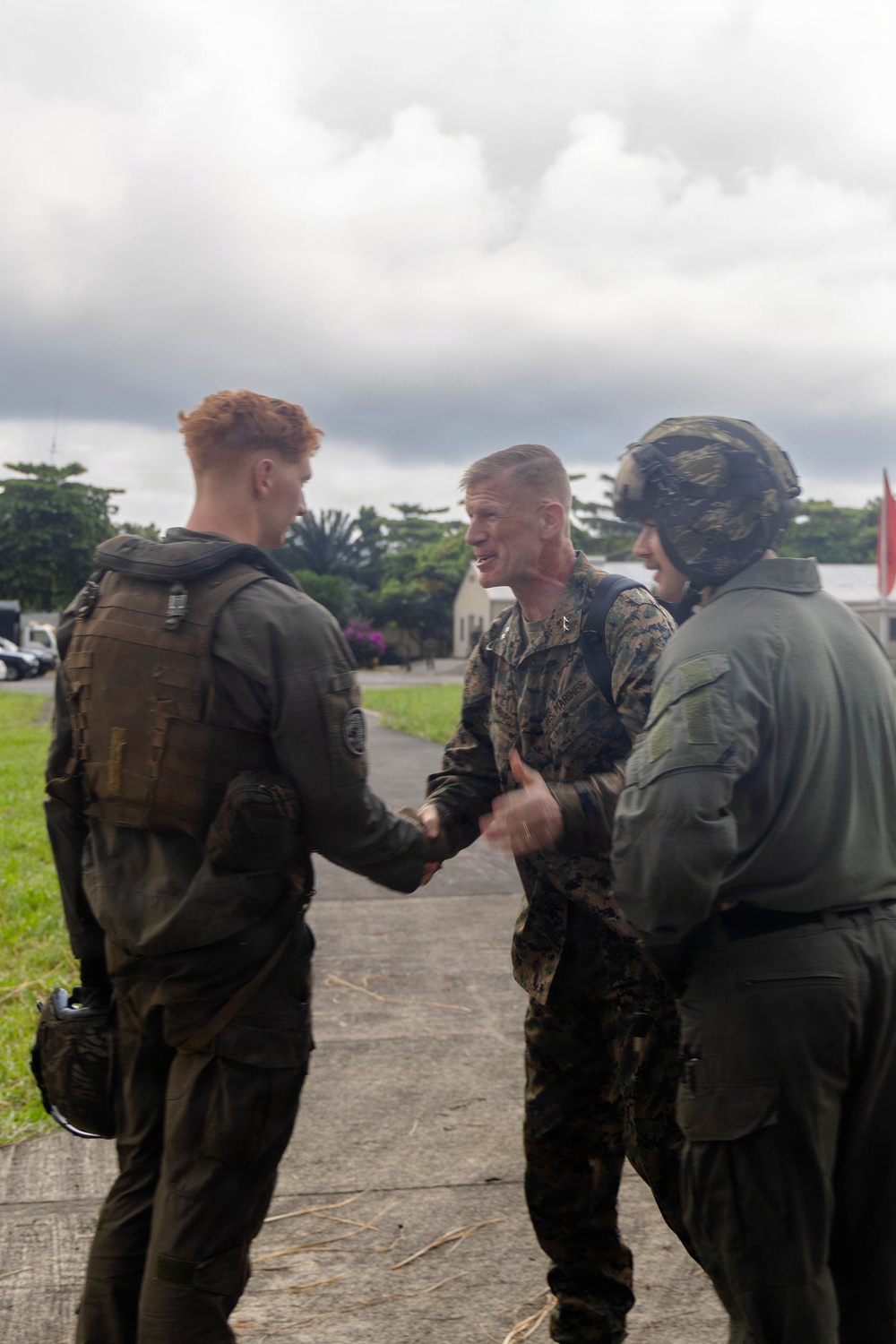 Three military personnel are engaged in a handshake interaction outdoors on a paved pathway. The individuals are wearing different military uniforms and gear, suggesting a multi-service or multinational context. The setting appears to be a tropical or subtropical military installation based on the vegetation and architecture visible in the background.