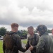 Three military personnel are engaged in a handshake interaction outdoors on a paved pathway. The individuals are wearing different military uniforms and gear, suggesting a multi-service or multinational context. The setting appears to be a tropical or subtropical military installation based on the vegetation and architecture visible in the background.