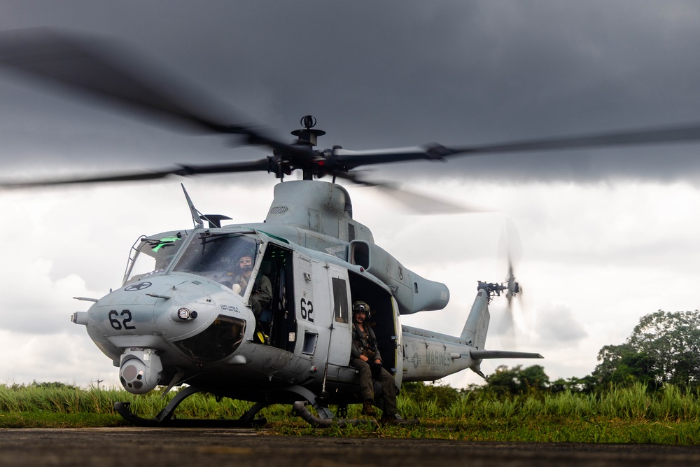 A U.S. Marines UH-1Y Venom helicopter (tail number 62) is on the ground with rotors spinning, indicating it is either preparing for takeoff or has just landed. A crew member in flight gear is seated in the open side door, while at least one pilot is visible in the cockpit. The setting appears to be a tropical or subtropical environment with lush green vegetation and overcast skies.