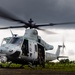 A U.S. Marines UH-1Y Venom helicopter (tail number 62) is on the ground with rotors spinning, indicating it is either preparing for takeoff or has just landed. A crew member in flight gear is seated in the open side door, while at least one pilot is visible in the cockpit. The setting appears to be a tropical or subtropical environment with lush green vegetation and overcast skies.