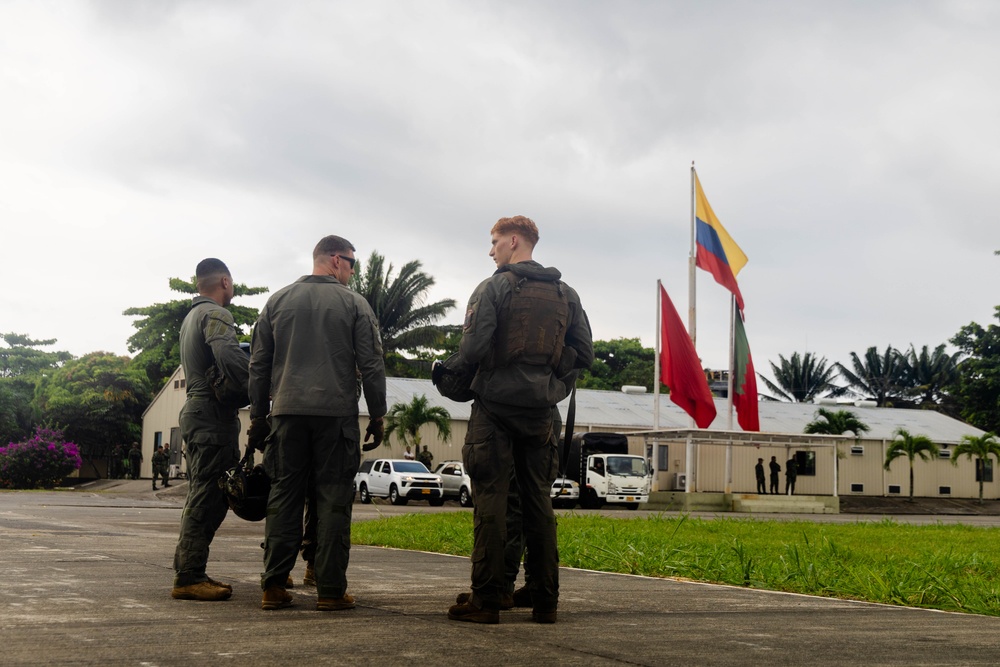 Three military personnel in olive/dark green flight suits are standing on a tarmac or paved surface, appearing to be in conversation. In the background, multiple flags are visible including the Colombian national flag (yellow, blue, red horizontal stripes), along with red and green flags on separate poles. The setting appears to be a military installation in a tropical environment, consistent with Colombia.