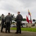 Three military personnel in olive/dark green flight suits are standing on a tarmac or paved surface, appearing to be in conversation. In the background, multiple flags are visible including the Colombian national flag (yellow, blue, red horizontal stripes), along with red and green flags on separate poles. The setting appears to be a military installation in a tropical environment, consistent with Colombia.