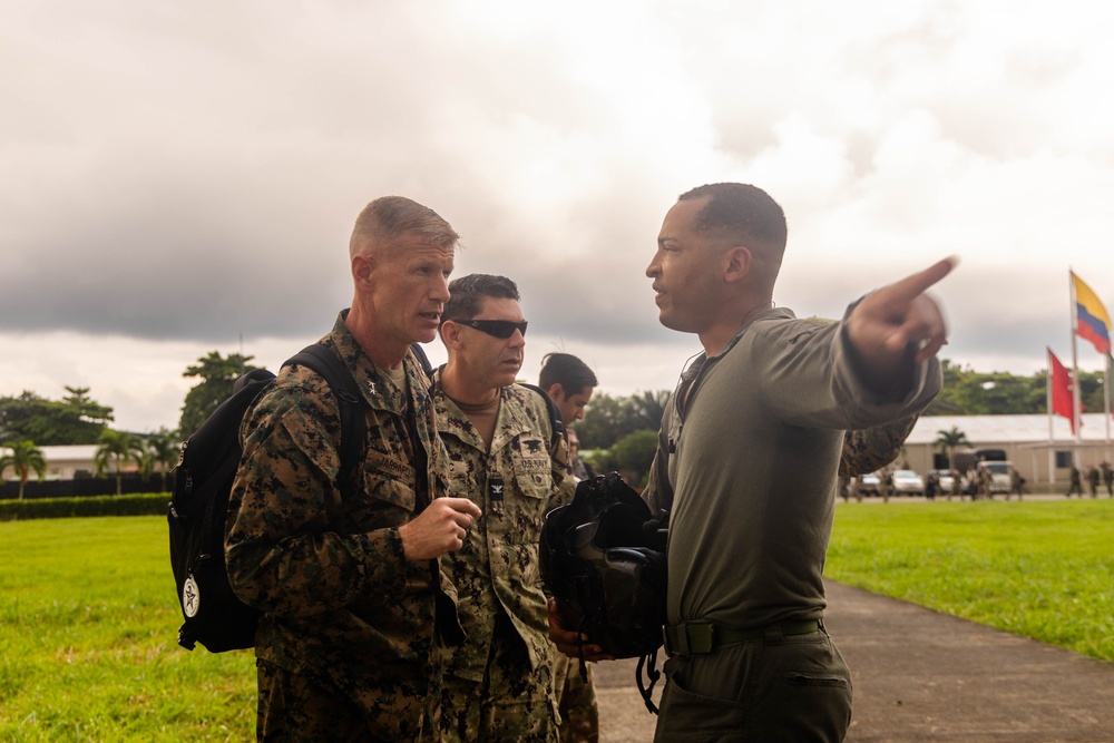 Three military personnel in uniform are engaged in a briefing or orientation outdoors on a military installation. One individual in olive drab uniform is pointing in a direction, apparently providing guidance or directions to two others in camouflage uniforms. Flags visible in the background include what appears to be the Colombian flag, suggesting this may be a joint military exercise or visit in Colombia.