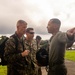 Three military personnel in uniform are engaged in a briefing or orientation outdoors on a military installation. One individual in olive drab uniform is pointing in a direction, apparently providing guidance or directions to two others in camouflage uniforms. Flags visible in the background include what appears to be the Colombian flag, suggesting this may be a joint military exercise or visit in Colombia.