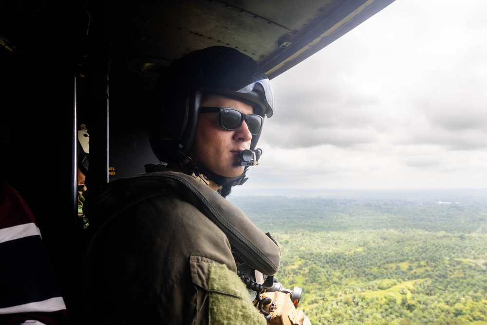 A military or law enforcement aircrew member is seated inside a helicopter, looking out over a vast green forested landscape below. The individual is wearing a flight helmet with a communications microphone, sunglasses, and tactical gear. The aircraft appears to be in flight at moderate altitude under an overcast sky.
