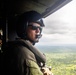 A military or law enforcement aircrew member is seated inside a helicopter, looking out over a vast green forested landscape below. The individual is wearing a flight helmet with a communications microphone, sunglasses, and tactical gear. The aircraft appears to be in flight at moderate altitude under an overcast sky.