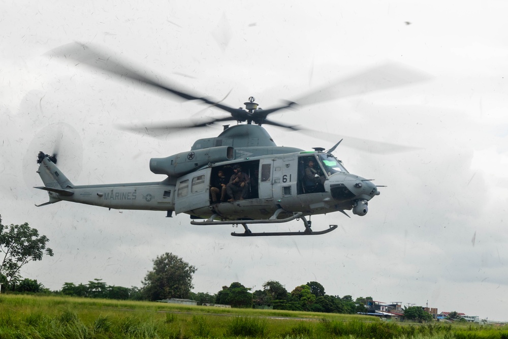 A U.S. Marines UH-1Y Venom helicopter (tail number 61) is captured in low-altitude flight over a grassy field in what appears to be a tropical environment. The helicopter's side door is open with at least one crew member visible in the cabin. The image was likely taken through a rain-spotted window, giving the photo a slightly obscured quality.