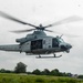 A U.S. Marines UH-1Y Venom helicopter (tail number 61) is captured in low-altitude flight over a grassy field in what appears to be a tropical environment. The helicopter's side door is open with at least one crew member visible in the cabin. The image was likely taken through a rain-spotted window, giving the photo a slightly obscured quality.