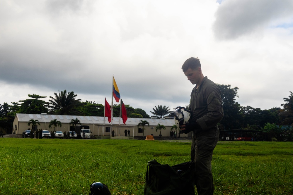 A military personnel member in olive/green flight suit stands on a grass field, holding a white flight helmet and examining equipment. In the background, multiple flags are visible including what appears to be the Colombian flag (yellow, blue, red) and red military/unit flags, alongside a low military installation building. The setting appears to be a military base in a tropical environment.