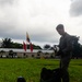 A military personnel member in olive/green flight suit stands on a grass field, holding a white flight helmet and examining equipment. In the background, multiple flags are visible including what appears to be the Colombian flag (yellow, blue, red) and red military/unit flags, alongside a low military installation building. The setting appears to be a military base in a tropical environment.