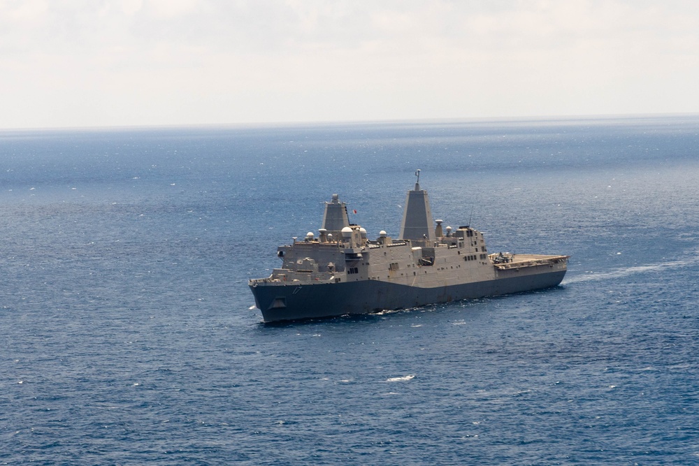 A large gray military naval vessel, consistent with a San Antonio-class amphibious transport dock (LPD) of the United States Navy, is underway on open blue ocean waters. The ship is photographed from an elevated aerial perspective, showing its full profile with a flat flight deck aft section and prominent superstructure with two masts. Sea conditions appear calm with light overcast skies.