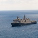 A large gray military naval vessel, consistent with a San Antonio-class amphibious transport dock (LPD) of the United States Navy, is underway on open blue ocean waters. The ship is photographed from an elevated aerial perspective, showing its full profile with a flat flight deck aft section and prominent superstructure with two masts. Sea conditions appear calm with light overcast skies.