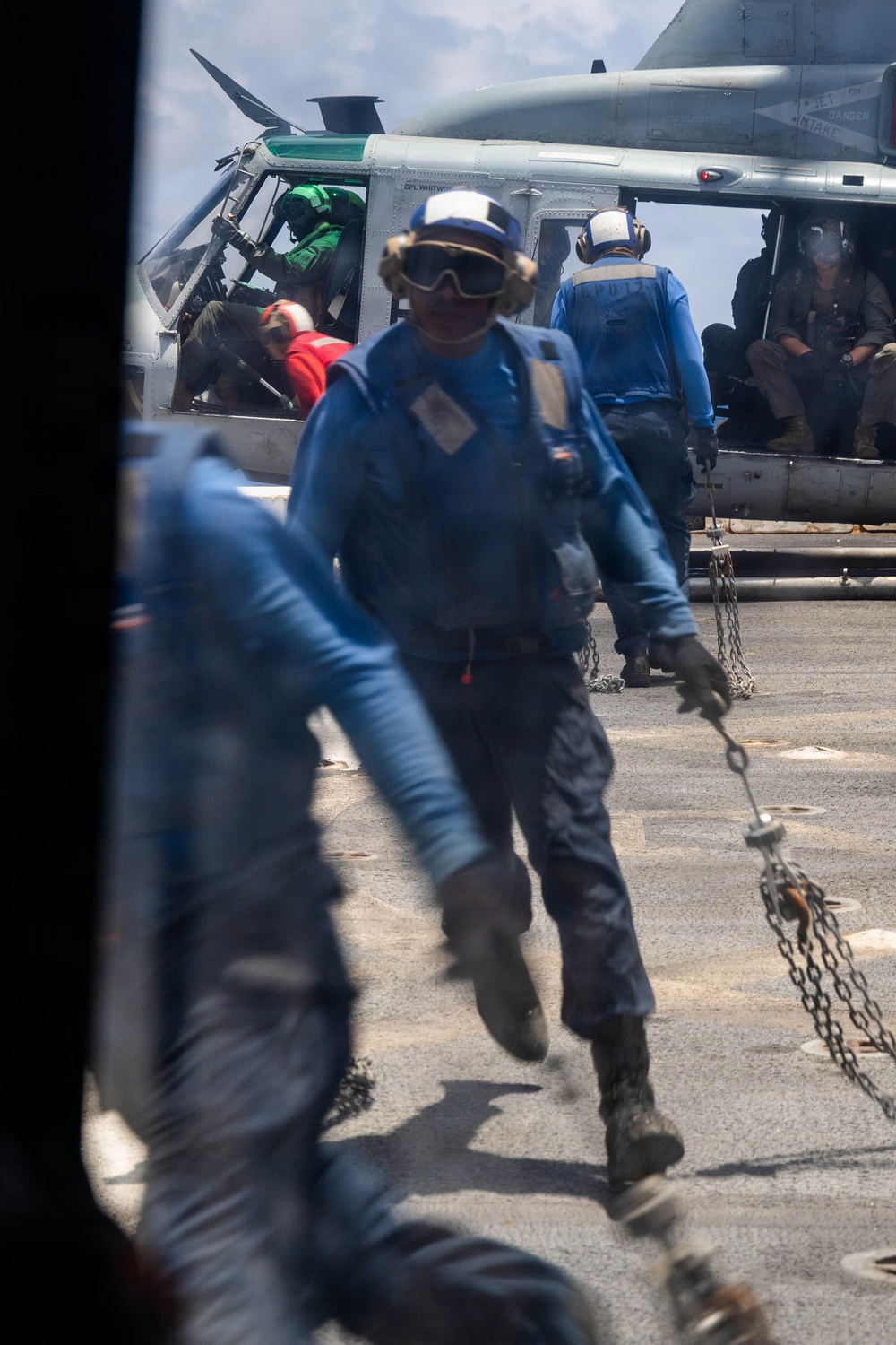The image shows naval flight deck crew members in blue jerseys working around a military helicopter on what appears to be a ship's flight deck. Personnel are actively moving and handling chains, likely securing or unsecuring the aircraft. The scene depicts standard carrier deck operations with multiple crew members in various colored jerseys performing assigned roles.