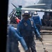 The image shows naval flight deck crew members in blue jerseys working around a military helicopter on what appears to be a ship's flight deck. Personnel are actively moving and handling chains, likely securing or unsecuring the aircraft. The scene depicts standard carrier deck operations with multiple crew members in various colored jerseys performing assigned roles.
