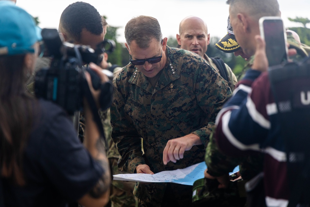 A senior military officer in U.S. Marine Corps digital camouflage uniform is reviewing a map or document outdoors, surrounded by a group of people including media personnel with cameras. The officer appears to be pointing at or examining specific details on the document. The scene suggests a field briefing or site visit with press coverage.