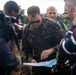 A senior military officer in U.S. Marine Corps digital camouflage uniform is reviewing a map or document outdoors, surrounded by a group of people including media personnel with cameras. The officer appears to be pointing at or examining specific details on the document. The scene suggests a field briefing or site visit with press coverage.