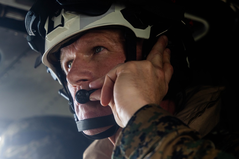 A close-up photograph of a military personnel wearing a white/tan helmet and MARPAT (Marine pattern) camouflage uniform. The individual is adjusting or securing a communications headset/microphone near their mouth. The image is taken from a low angle in what appears to be a dimly lit interior environment, likely a military vehicle or aircraft.