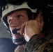 A close-up photograph of a military personnel wearing a white/tan helmet and MARPAT (Marine pattern) camouflage uniform. The individual is adjusting or securing a communications headset/microphone near their mouth. The image is taken from a low angle in what appears to be a dimly lit interior environment, likely a military vehicle or aircraft.