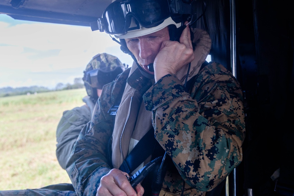 A military personnel wearing MARPAT (Marine pattern) camouflage uniform and a white flight helmet with attached goggles is seated inside what appears to be a military helicopter. The individual is adjusting a communication headset or microphone near their mouth. A second person in similar military attire is visible in the background.