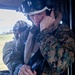 A military personnel wearing MARPAT (Marine pattern) camouflage uniform and a white flight helmet with attached goggles is seated inside what appears to be a military helicopter. The individual is adjusting a communication headset or microphone near their mouth. A second person in similar military attire is visible in the background.