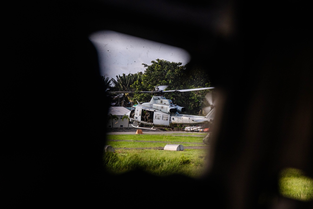 A military helicopter, appearing to be a U.S. Marine Corps UH-1Y Venom or similar utility helicopter, is photographed through a gap or hole in a dark foreground structure. The aircraft is on or near a grassy landing area surrounded by tropical vegetation. The framing technique creates a voyeuristic or surveillance-like perspective.