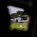 A military helicopter, appearing to be a U.S. Marine Corps UH-1Y Venom or similar utility helicopter, is photographed through a gap or hole in a dark foreground structure. The aircraft is on or near a grassy landing area surrounded by tropical vegetation. The framing technique creates a voyeuristic or surveillance-like perspective.