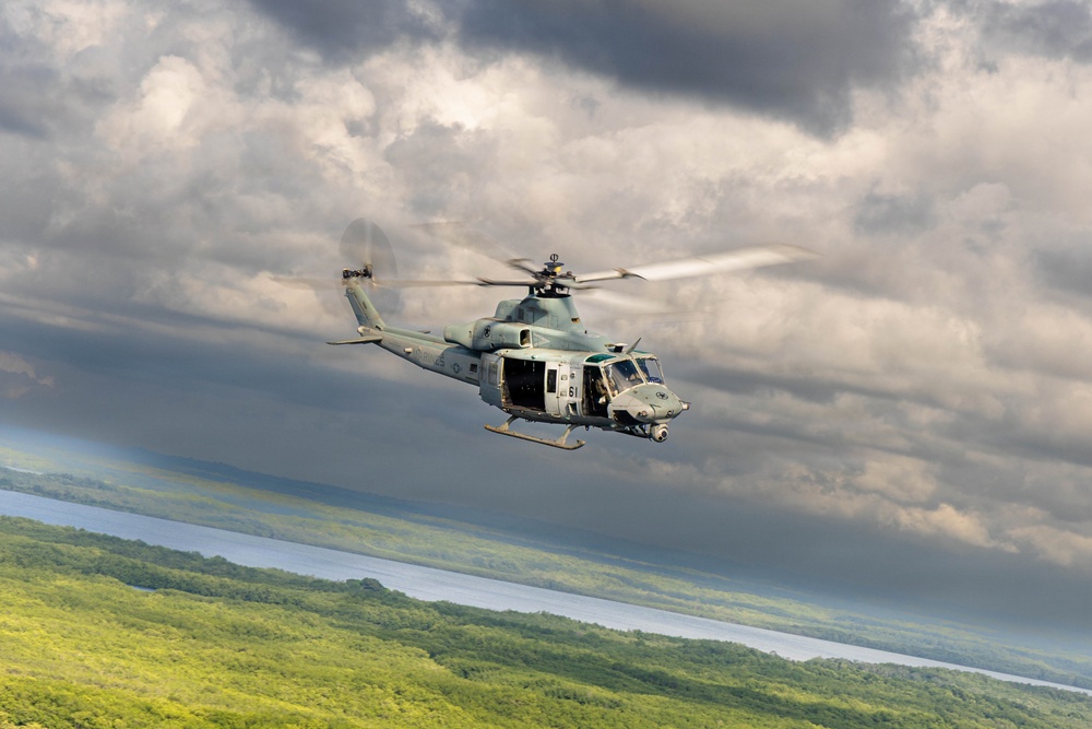 A military helicopter, appearing to be a Bell UH-1Y Venom (Super Huey), is captured in flight over a lush green forested landscape with a body of water visible in the background. The aircraft is painted in a light gray military scheme and has its side door open. The image is taken from another airborne platform, suggesting an air-to-air photography mission.